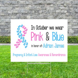 Pode incluir: Uma placa branca em um suporte de metal com texto e uma fita feita de pegadas rosa e azuis. A placa diz: "Em outubro vestimos rosa e azul em homenagem a: Adrian James. Conscientiza&ccedil;&atilde;o e lembran&ccedil;a da perda gestacional e infantil."