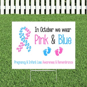 Pode incluir: Placa de jardim branca com o texto "In October we wear Pink & Blue" e "Pregnancy & Infant Loss Awareness & Remembrance". A placa apresenta uma fita rosa e azul feita de pegadas.