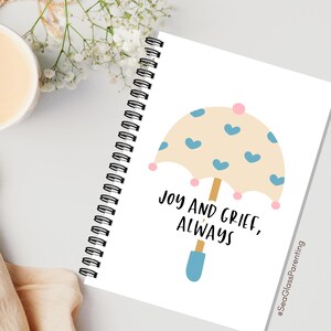 May include: A white spiral-bound notebook with a cream-coloured cover. The cover features a cartoon umbrella design with blue hearts and the text "Joy and Grief, Always". The notebook is on a white surface.