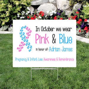 Pode incluir: Uma placa de jardim branca com texto e uma fita feita de pegadas rosa e azuis. A placa diz: "Em outubro vestimos rosa e azul em homenagem a Adrian James. Conscientiza&ccedil;&atilde;o e lembran&ccedil;a da perda gestacional e infantil."