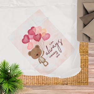 Könnte beinhalten: Eine weiche, hellrosa Decke mit einem Teddybären, der herzförmige Ballons in Rosa- und Pfirsichtönen hält. Die Worte "I am always thinking of you" sind in einer Schriftart geschrieben. Die Decke liegt auf einer weißen Oberfläche.