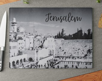 May include: A rectangular black and white cutting board featuring a detailed image of Jerusalem. The word "Jerusalem" is written in cursive across the top. The image shows buildings, arches, and a large wall with people gathered.