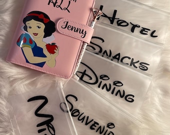 May include: A pink travel planner with the text "Fairest of them all" and "Jenny" on the cover. The planner is surrounded by clear plastic bags with the words "Tickets", "Hotel", "Snacks", "Dining", "Souvenirs", and "Misc." printed on them.