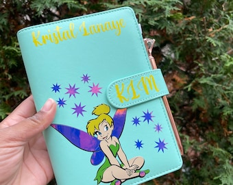 May include: A light blue journal with a cartoon image of Tinkerbell and star accents. The journal has the name "Kristal Lunaye" in gold lettering on the top and the initials "KIM" on the clasp. A rose gold pen is attached to the side.
