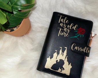 May include: Black leather planner with gold glitter accents. The planner features a silhouette of Belle and the Beast in front of the castle from Beauty and the Beast. The text "Tale as old as Time" is written in gold script. The planner is personalized with the name "Carrillo".