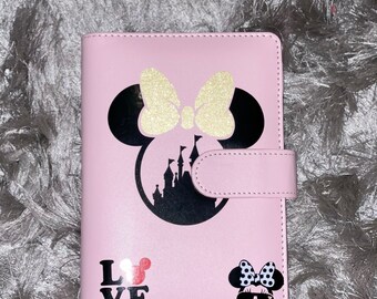 May include: A pink leatherette notebook with a black and gold Mickey Mouse design. The notebook has a black castle silhouette, a gold bow, and the word "LOVE" with a Mickey Mouse head.