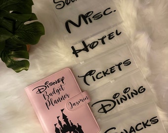 May include: Pink Disney budget planner with a castle and Mickey Mouse silhouette. The planner has a clear plastic pocket on the front with the word "Jasmin" written on it. The planner is surrounded by clear plastic envelopes with the words "Souvenirs", "Misc", "Hotel", "Tickets", "Dining", and "Snacks" written on them.