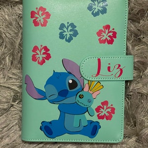 May include: A light blue and green faux leather journal featuring a cartoon Stitch character holding a doll. The journal has a flap closure with the name "Liz" written on it. The journal is decorated with pink and blue hibiscus flowers.