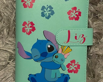 May include: A light blue and green faux leather journal with a cartoon Stitch character holding a doll. The journal has a flap closure with the name "Liz" written on it. The journal is decorated with pink and blue hibiscus flowers.