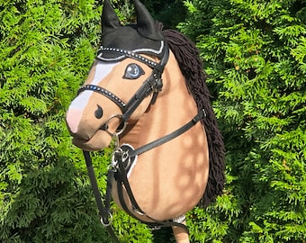 Puede incluir: Un caballo de juguete marrón con crin, orejas y brida negras. La brida está adornada con tachuelas plateadas. El caballo tiene una raya blanca en la cara y está sobre un fondo de follaje verde.