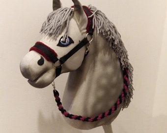 Puede incluir: Una cabeza de caballo de juguete de color gris, con una brida negra y burdeos y una rienda trenzada. El caballo tiene ojos azules y pelo de hilo gris. El palo es de madera.