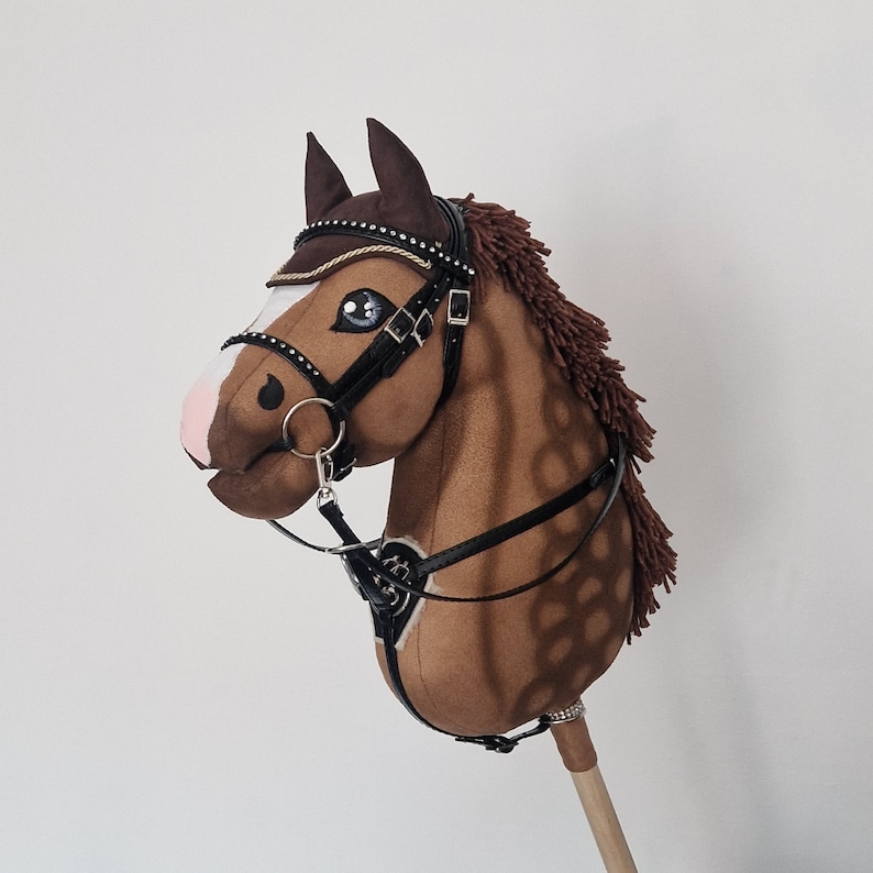 Pu&ograve; includere: Un cavallo a dondolo marrone con briglia e redini nere. Il cavallo ha una criniera marrone e un manico di legno. Il viso del cavallo &egrave; dettagliato con occhi blu, naso rosa e segni bianchi.