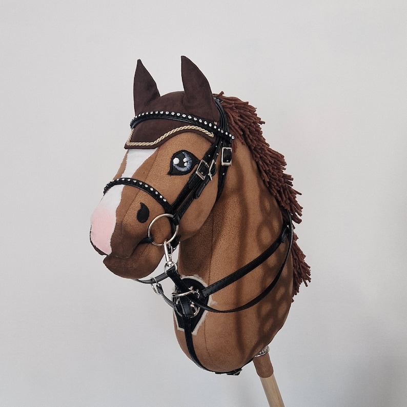 Pu&ograve; includere: Un cavallo a dondolo marrone e bianco con briglia e redini nere. Il cavallo ha criniera e orecchie marroni e occhi blu. La briglia &egrave; decorata con strass. Il cavallo &egrave; montato su un bastone di legno.