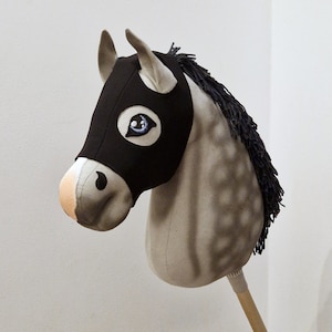 Op de afbeelding: Een grijs en wit pluche paardenhoofd met een zwart stoffen masker dat zijn gezicht bedekt. Het paard heeft een blauw oog en een zwarte manen. Het paard is bevestigd aan een houten stok.