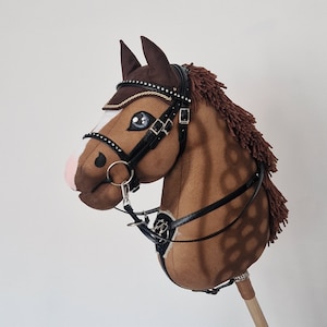 Pu&ograve; includere: Un cavallo a dondolo marrone con una briglia nera dettagliata, una criniera marrone e un bastone di legno. Il cavallo ha occhi blu, un naso rosa e una fascia frontale decorativa. Il bastone &egrave; lungo circa 76 cm.