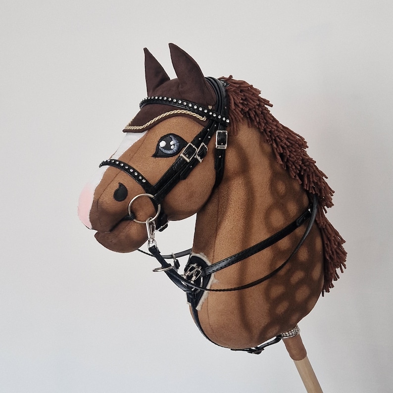 Pu&ograve; includere: Un cavallo a dondolo marrone con una briglia nera dettagliata, una criniera marrone e un bastone di legno. Il cavallo ha occhi blu, un naso rosa e una fascia frontale decorativa. Il bastone &egrave; lungo circa 76 cm.