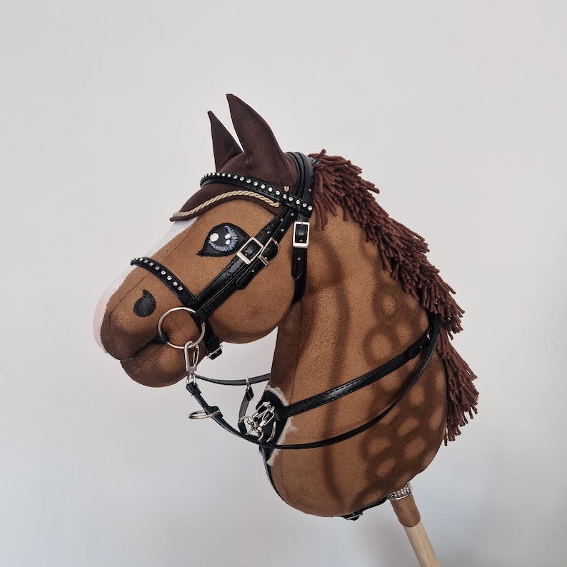 Pu&ograve; includere: Un cavallo a dondolo marrone con briglia e redini nere. Il cavallo ha criniera e orecchie marroni, con una striscia bianca sul muso. La briglia &egrave; decorata con strass. Il cavallo &egrave; montato su un bastone di legno.