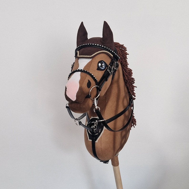 Pu&ograve; includere: Un cavallo a dondolo marrone con briglie e redini dettagliate. Il cavallo ha una macchia bianca sul muso e un naso rosa. Le briglie sono nere con dettagli color argento. La criniera &egrave; di un marrone pi&ugrave; scuro.