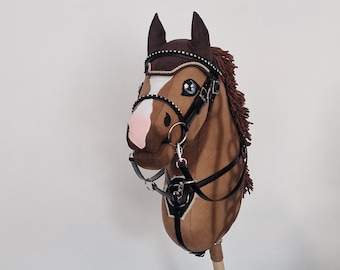 Puede incluir: Un caballo de juguete marrón con brida y riendas detalladas. El caballo tiene una mancha blanca, hocico rosa y ojos azules. La melena es de color marrón oscuro y el palo es de madera.