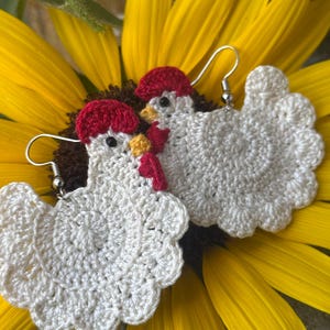 May include: A pair of handmade crochet chicken earrings. The earrings are white with red combs and yellow beaks. The earrings are displayed on a yellow sunflower.