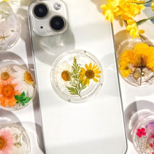 Könnte beinhalten: Eine weiße Handyhülle mit einem durchsichtigen Handygriff mit gepressten gelben und weißen Blumen. Der Handygriff ist ein beliebtes Accessoire für Handys.