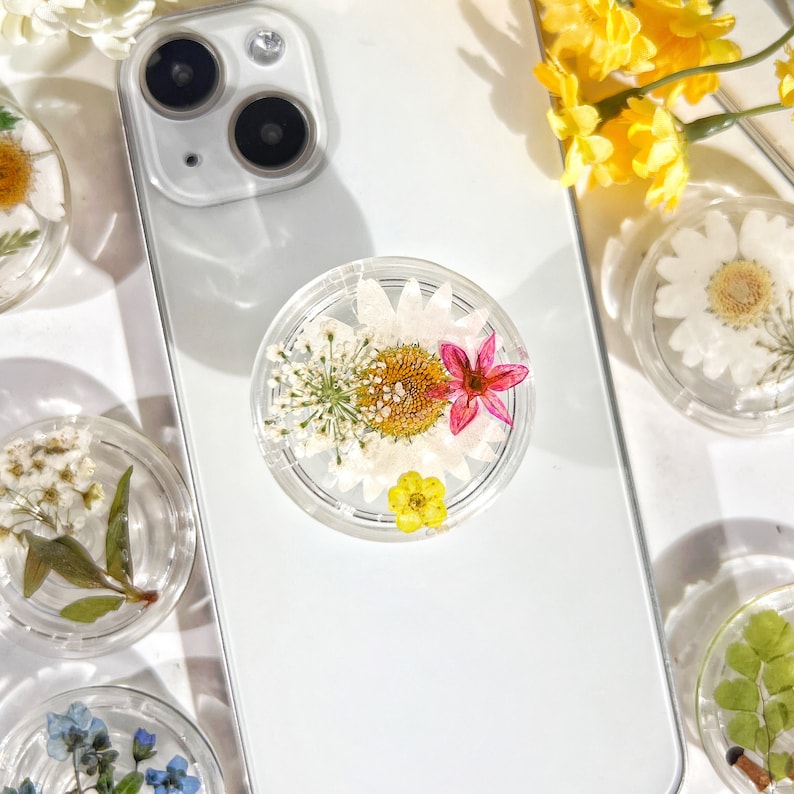 May include: A white phone case with a clear phone grip featuring pressed flowers. The flowers are white, pink, and yellow. The phone case is white and has a silver camera lens.
