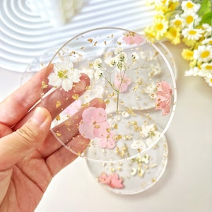 May include: Set of three clear resin coasters with pressed flowers and gold leaf. The coasters feature pink and white flowers, and gold leaf flakes.
