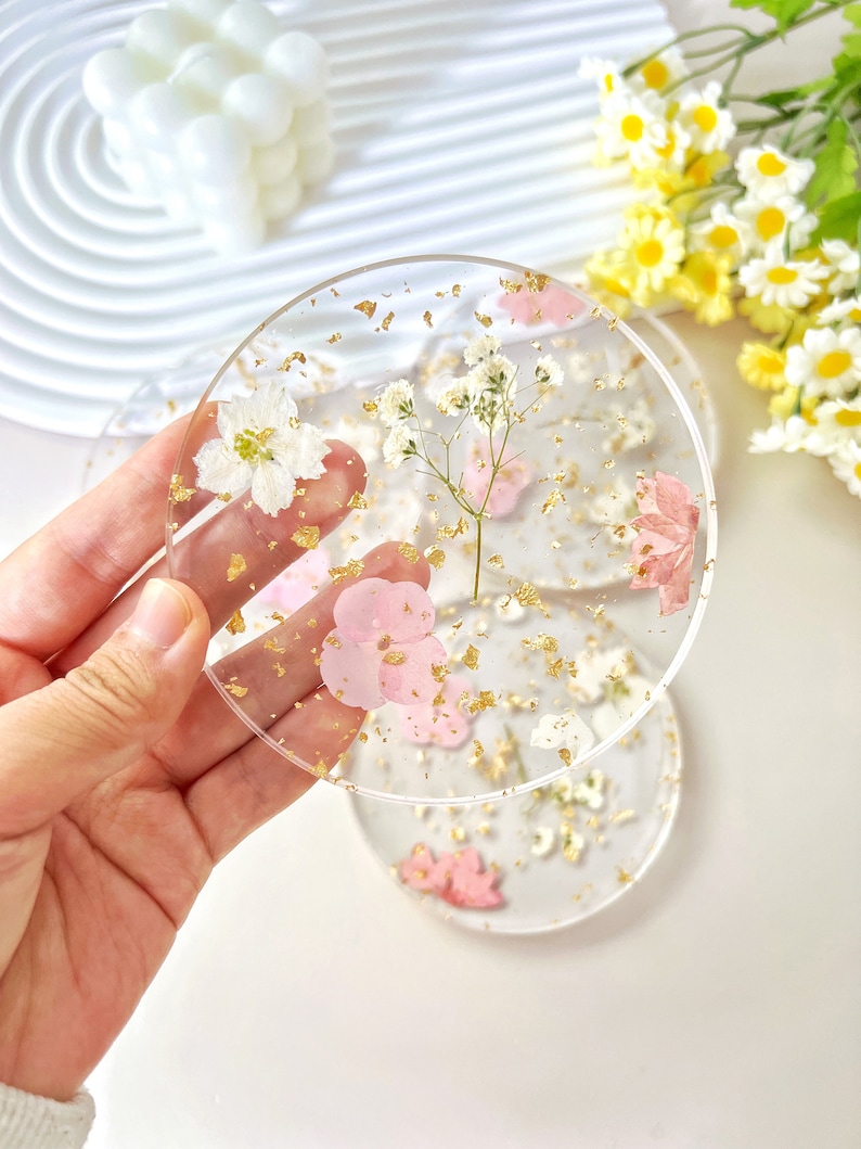 May include: Set of three clear resin coasters with pressed flowers and gold leaf. The coasters feature pink and white flowers, and gold leaf flakes.