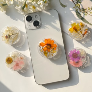 Puede incluir: Cuatro agarres para teléfono con flores prensadas. Los agarres son transparentes y muestran las flores con detalle. Las flores son naranja, amarillo, rosa y morado.