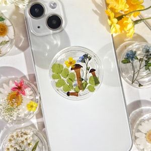 May include: A white phone case with a clear phone grip featuring pressed flowers and mushrooms. The phone grip is a popular accessory for phones.