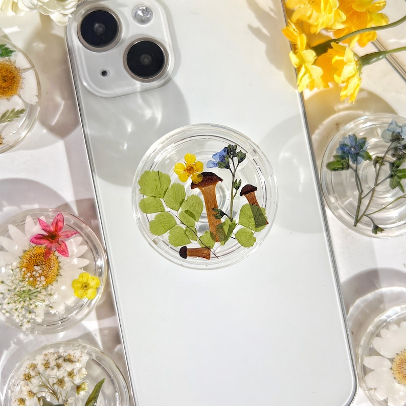 May include: A white phone case with a clear phone grip featuring pressed flowers and mushrooms. The phone grip is a popular accessory for phones.