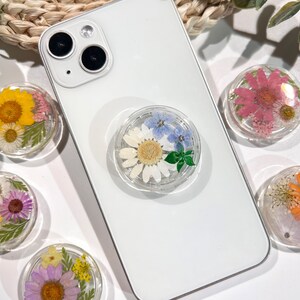 May include: Clear phone grip with a white daisy, blue flowers, and green leaves encased in resin, attached to a white phone. Several other floral resin grips with yellow, pink, and purple flowers are visible.