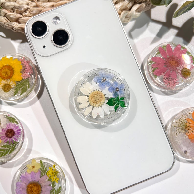 May include: Clear phone grip with a white daisy, blue flowers, and green leaves encased in resin, attached to a white phone. Several other floral resin grips with yellow, pink, and purple flowers are visible.