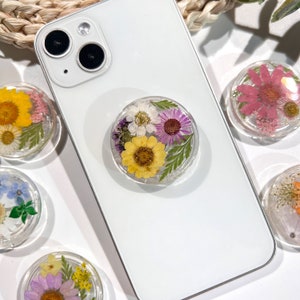 May include: A white smartphone with a clear phone grip featuring pressed flowers in shades of yellow, purple, and white. Several other floral phone grips are visible, each with different flower arrangements.