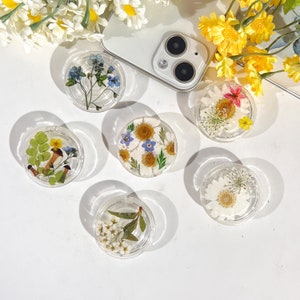 May include: Six clear resin phone grips with different pressed flower designs. The designs include white and blue flowers, white flowers with yellow centres, and a mix of white, yellow, and pink flowers.