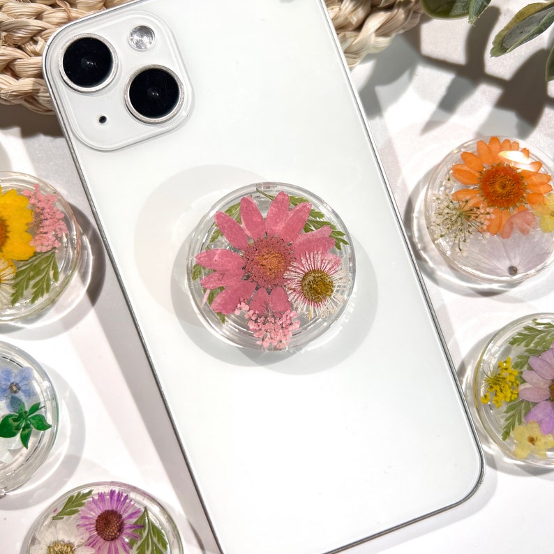 May include: Clear phone grip with a pink flower and other dried flowers encased in resin. The phone grip is attached to a white smartphone. Other phone grips with different colored flowers are also visible.