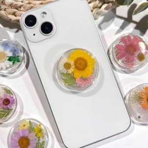 May include: Clear phone grip with a pressed yellow sunflower, white daisy, and pink flowers. The round grip is attached to a white smartphone. Other grips with different pressed flowers are visible.