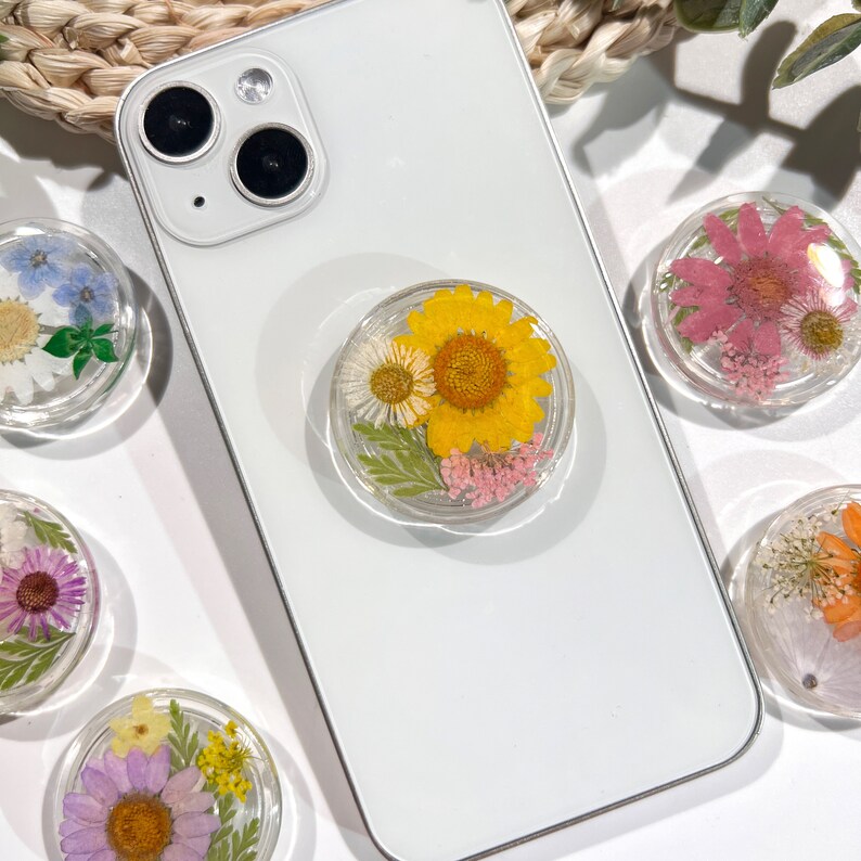 May include: Clear phone grip with a pressed yellow sunflower, white daisy, and pink flowers. The round grip is attached to a white smartphone. Other grips with different pressed flowers are visible.