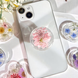 May include: A white iPhone with a clear phone grip featuring pressed pink and white flowers. The phone is lying on a white surface with other phone grips featuring pressed flowers in different colors.