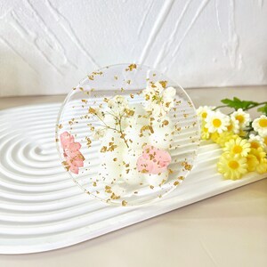 May include: A clear round coaster with dried flowers and gold flakes embedded in the resin. The coaster is on a white surface.