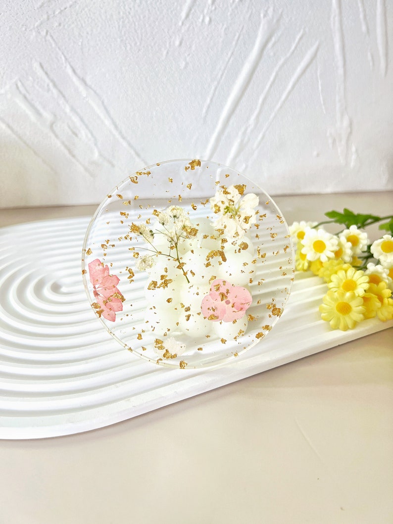 May include: A clear round coaster with dried flowers and gold flakes embedded in the resin. The coaster is on a white surface.