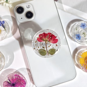 May include: A clear phone grip with a red flower and gold leaf accents. The grip is designed to be attached to the back of a phone.