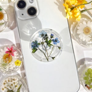 Puede incluir: Una agarradera de teléfono transparente con un diseño de flores prensadas. El diseño presenta flores azules de nomeolvides y hojas verdes.
