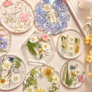 May include: Set of ten round and hexagonal resin coasters with pressed flowers and leaves. The coasters feature a variety of floral designs, including blue and white flowers, yellow and white flowers, and pink and white flowers.