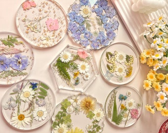 May include: Set of ten round and hexagonal resin coasters with pressed flowers and leaves. The coasters feature a variety of floral designs, including blue and white flowers, yellow and white flowers, and pink and white flowers.