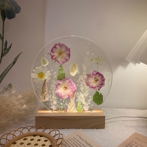 May include: A clear round resin lamp base with dried flowers and leaves encased in the resin. The lamp base is on a wooden stand. The lamp base is lit and the flowers are illuminated.