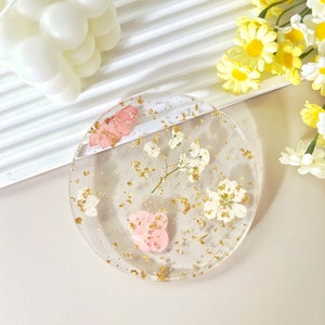 May include: A clear round coaster with gold flakes, dried white flowers, and a pink heart. The coaster is on a light beige surface.