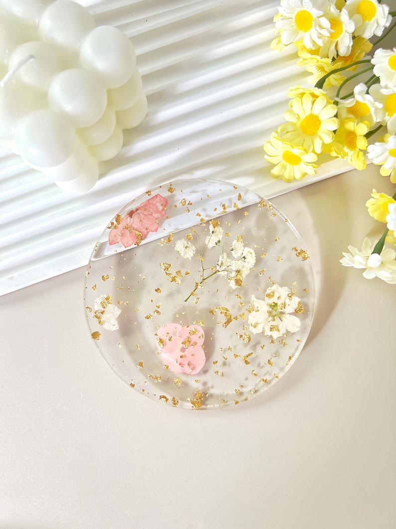 May include: A clear round coaster with gold flakes, dried white flowers, and a pink heart. The coaster is on a light beige surface.