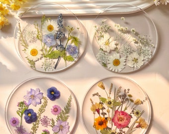 May include: Four round clear resin coasters with pressed flowers. The coasters feature different flower arrangements, including blue and purple flowers, white flowers, yellow and pink flowers, and a mix of white, yellow, and pink flowers.