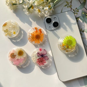 Op de afbeelding: Vijf verschillende telefoonbeugels met geperste bloemen erin. De beugels zijn helder en rond, en elk heeft een andere bloemenarrangement. De telefoonbeugels bevinden zich op een wit oppervlak.